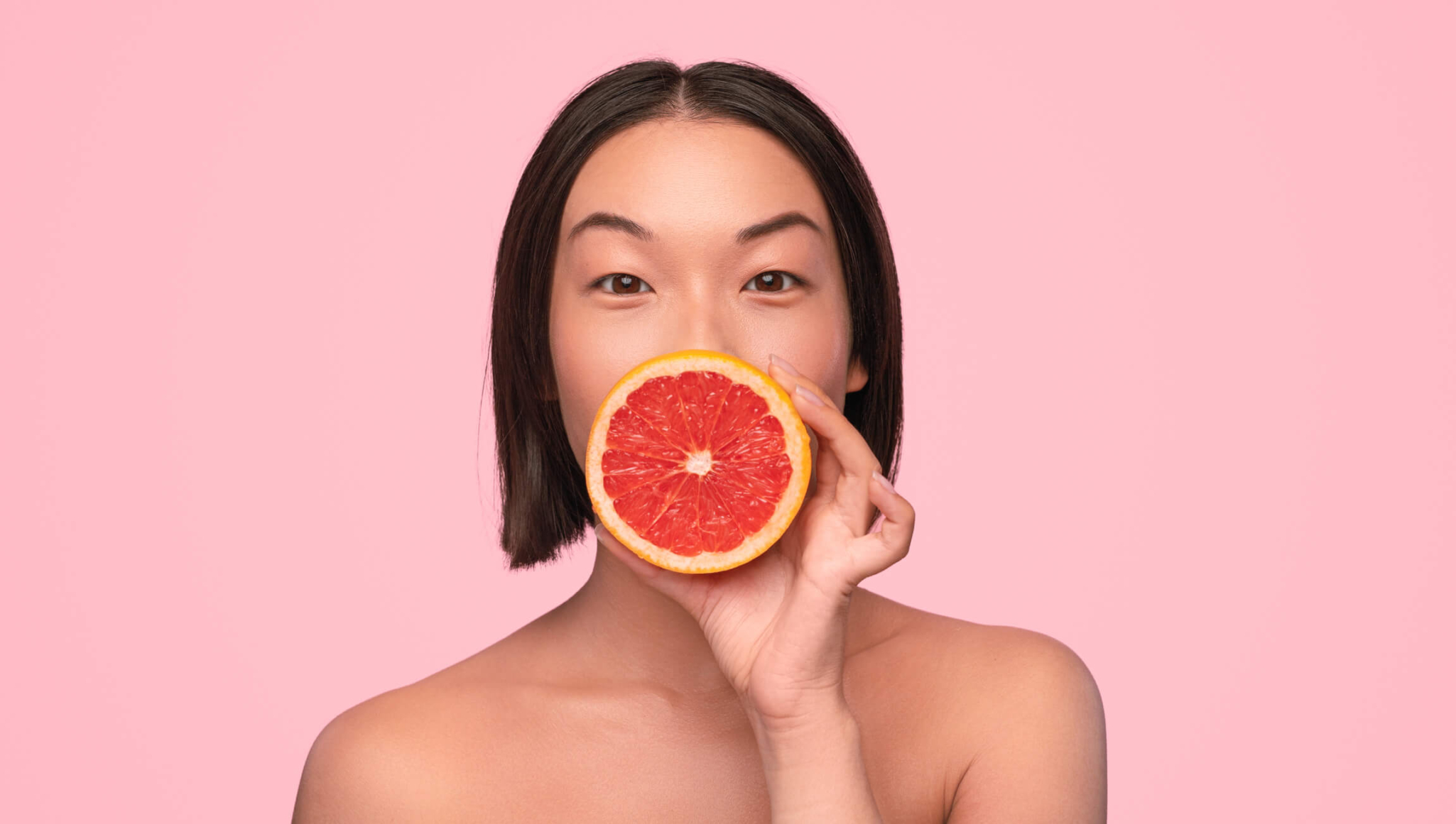 Young woman holding a red grapefruit over her face with a pink background.