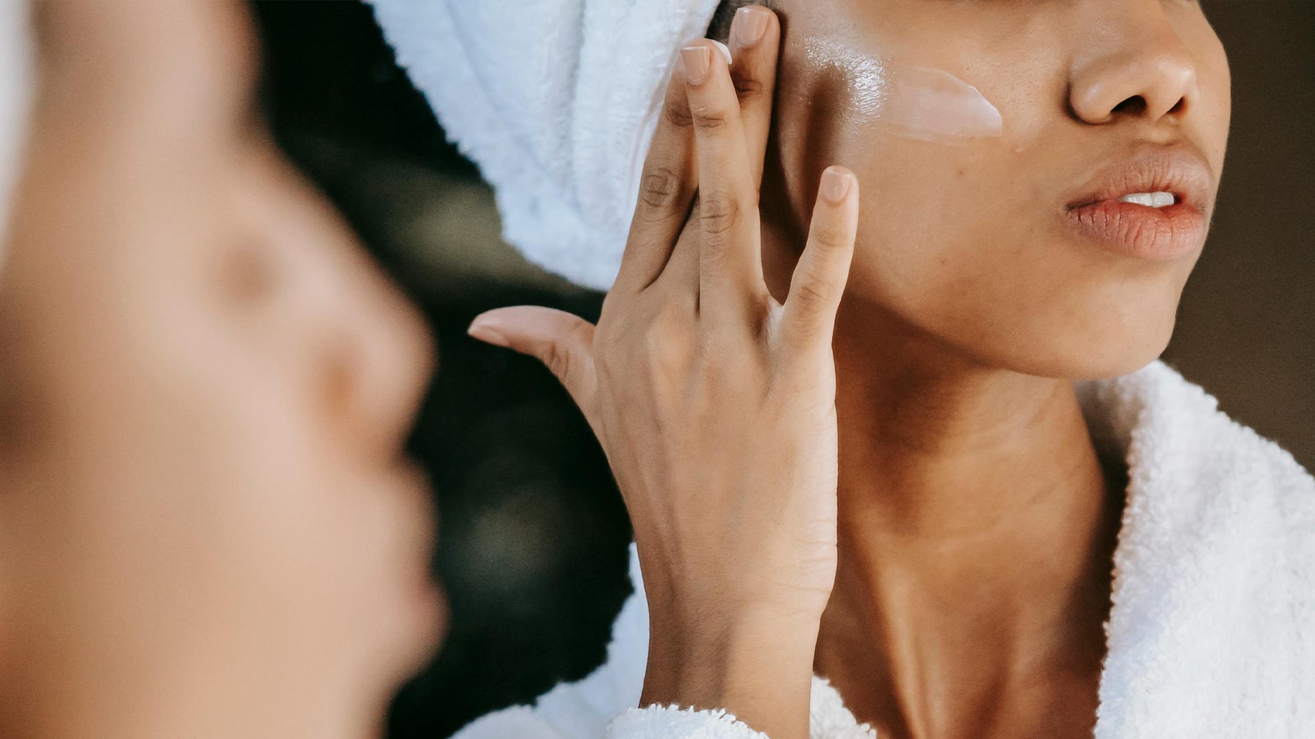 Shallow focus of woman in bathrobe applying moisturizer on her cheek
