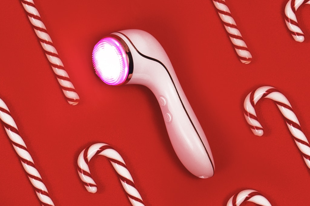 The reVive Light Therapy Lux Collection Sonique light therapy sonic cleanser is lying on its side with the lights on. It is on a red background with candy canes placed around it.