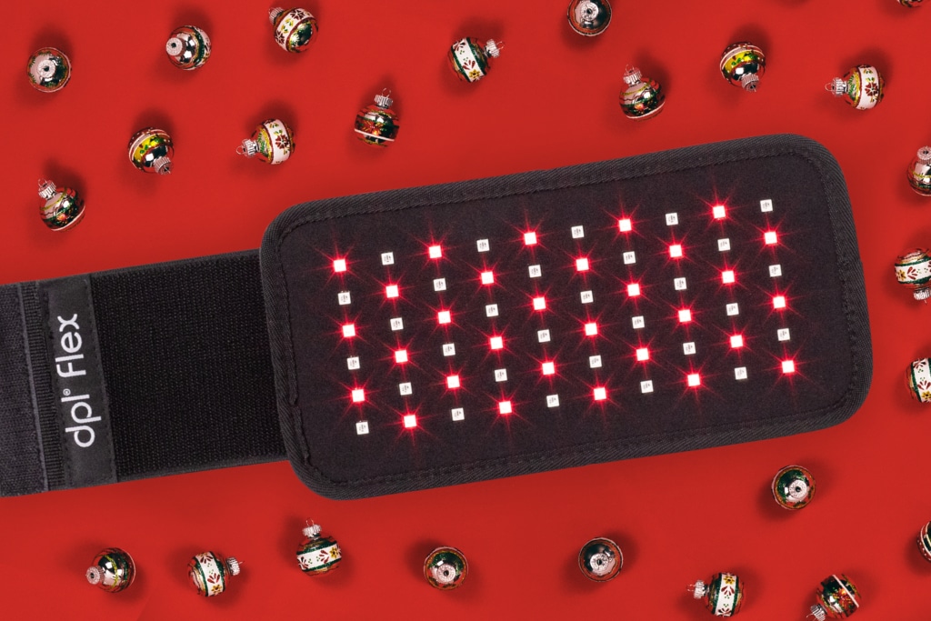 The dpl Flex Pad light therapy pain relief device is laying on a red background. Its lights are on, and there a tiny holiday ornaments scattered around it.