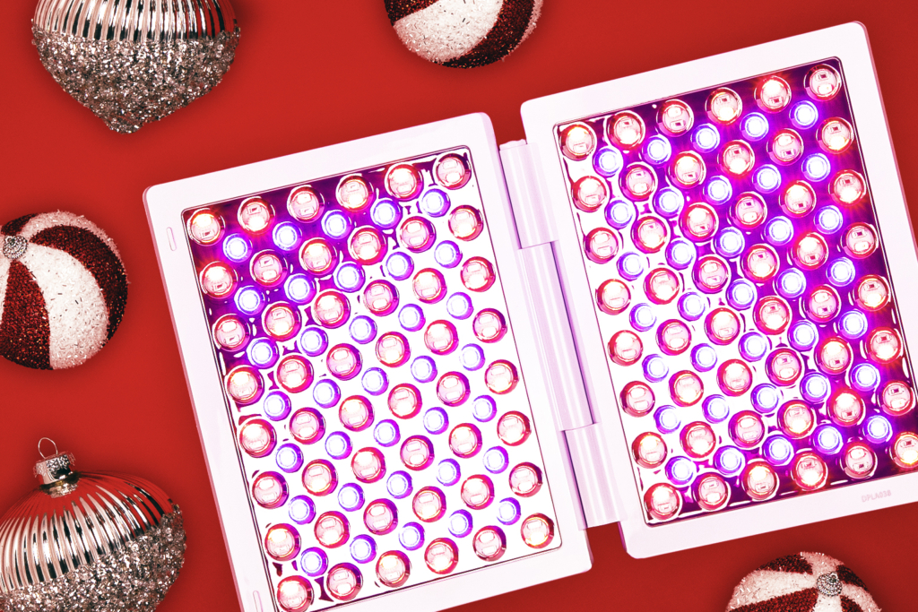 The dpl IIa Lux Collection LED Light Therapy treatment panel is lying open like a book with the lights on, giving it a purplish pink glow. It is on a red background, surrounded by holiday ornaments.