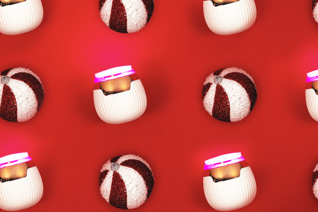 Several Lux Collection Sonique Mini light therapy cleansers are arranged in a pattern on a red background. Their lights are on, and they are in a checkerboard pattern with holiday ornaments.