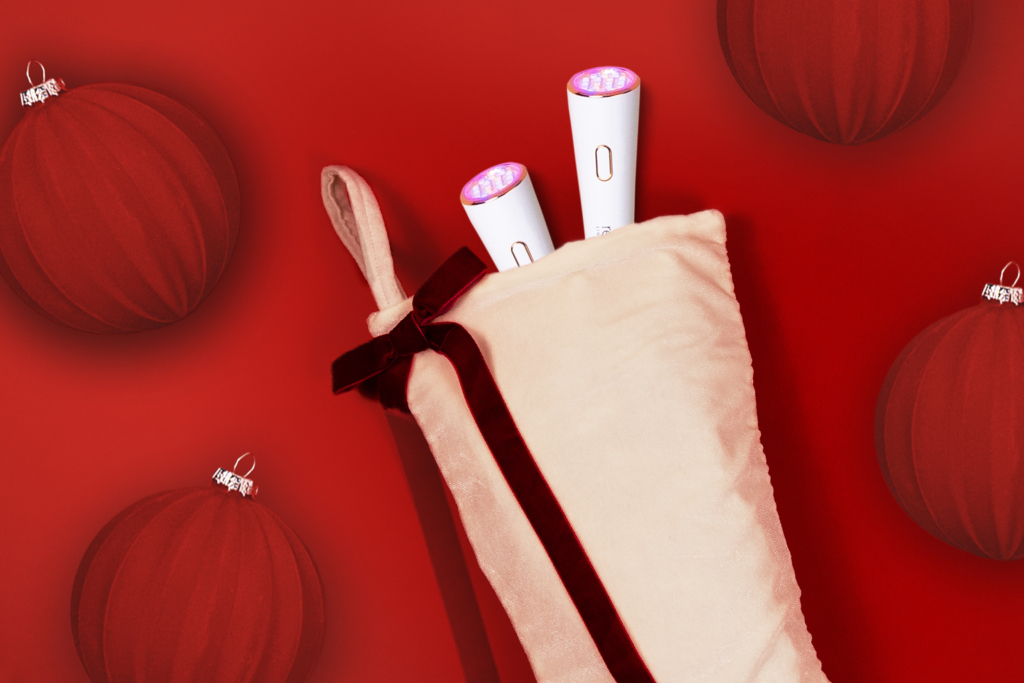 Two Lux Collection light therapy wands are sticking out of a white Christmas stocking. The stocking is on a red background surrounded by red velvety ornaments.