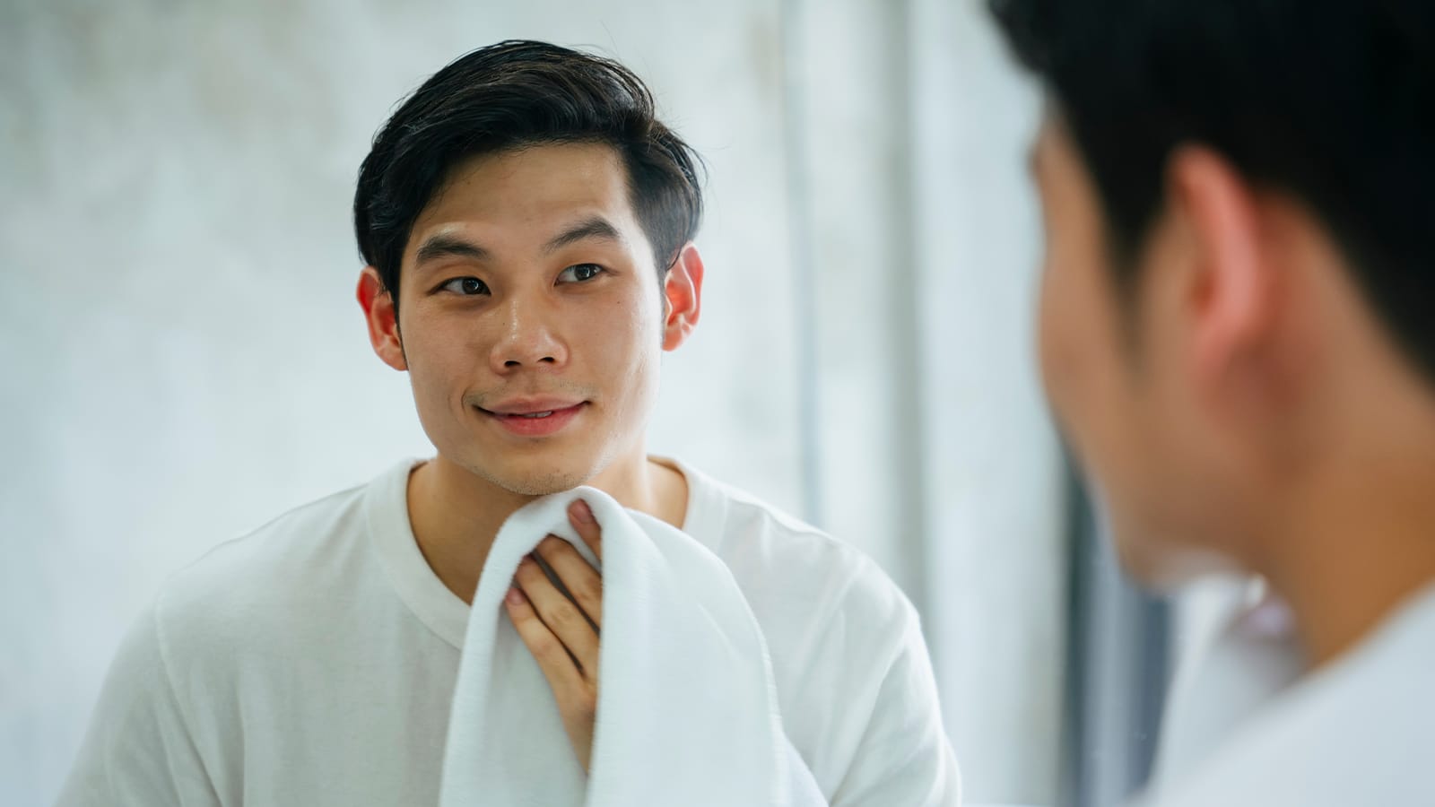 young man smiling in the mirror as he finishes washing his face with a white towel