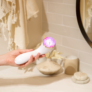 Lux Collection Sonique sonic cleanser in the hands of a woman in front of a bathroom counter; the LED lights are on and the serum massage brush head is facing upwards