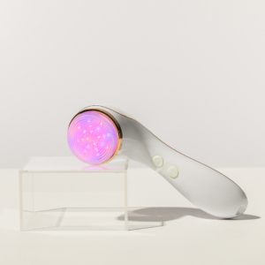 Lux Collection Sonique sonic cleanser rests on clear acrylic block, with LED lights on and cleansing brush head facing the viewer