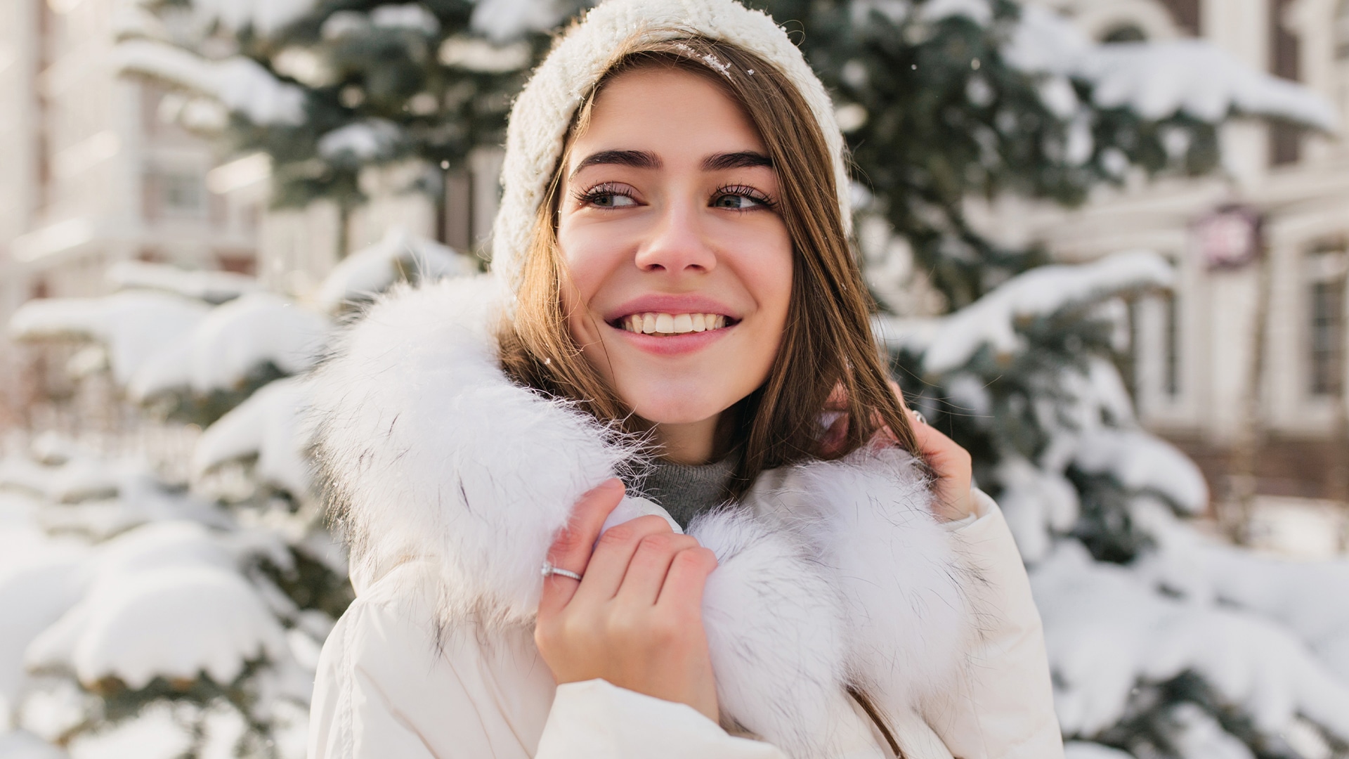 Woman wearing white winter wear smiling in front of a backdrop of snowy trees