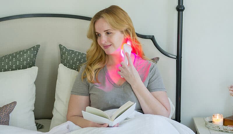 woman reading in bed while using red light therapy to reduce wrinkles on her neck