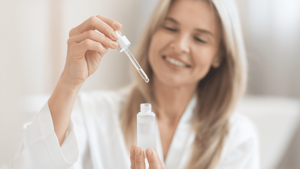 Woman in bathrobe removes dropper holding clear skincare serum from a frosted glass bottle 